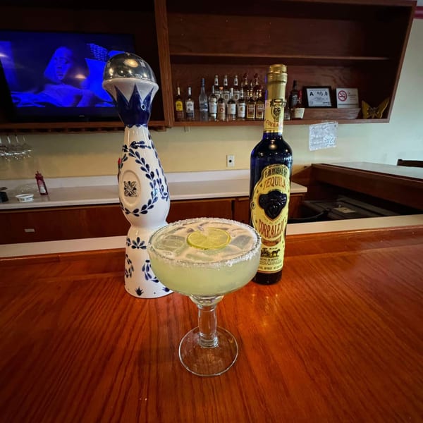 Margarita with lime and salt rim on a wooden bar next to tequila bottles and a TV.