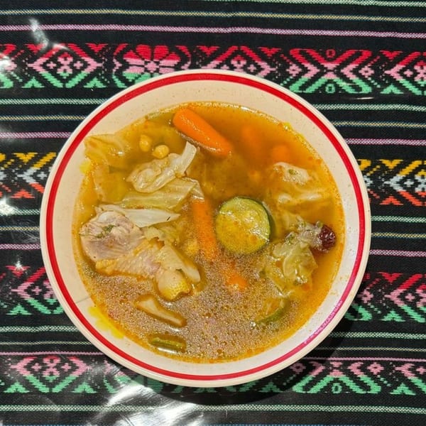 Hearty Mexican soup with chicken, carrots, zucchini, and cabbage in a white bowl with red trim.