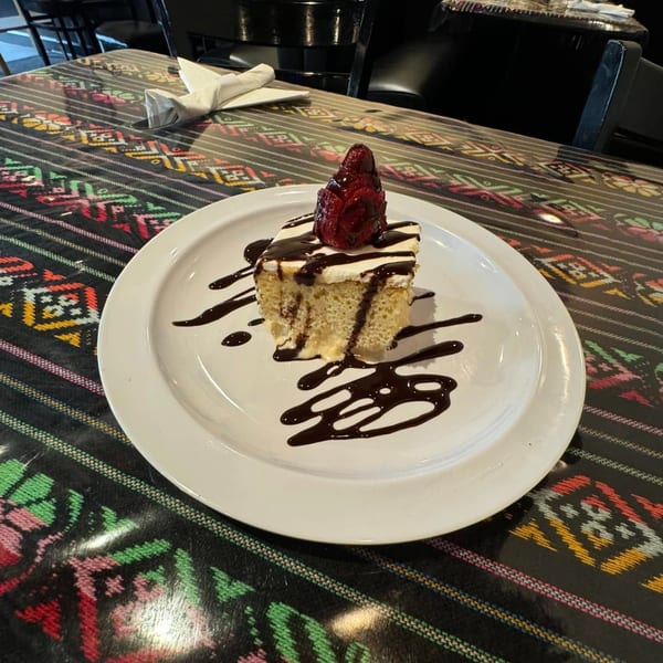 Slice of tres leches cake with whipped cream, chocolate drizzle, and a strawberry on a colorful patterned tablecloth.