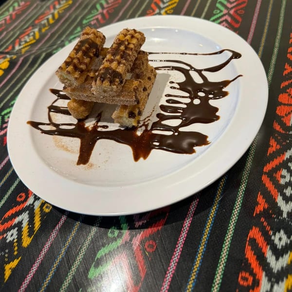 Churros drizzled with chocolate sauce and cinnamon sugar on a white plate, on a colorful patterned tablecloth.